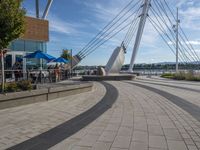 a sidewalk by the water with people walking and sitting on tables near a boat sculpture