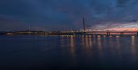 a sail boat sits at dusk near a large bridge in the water, lights shine bright