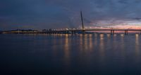 a sail boat sits at dusk near a large bridge in the water, lights shine bright