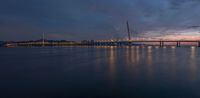 a sail boat sits at dusk near a large bridge in the water, lights shine bright