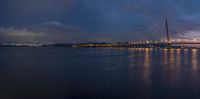 a sail boat sits at dusk near a large bridge in the water, lights shine bright