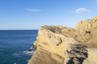 two people stand on the edge of a cliff next to the ocean in a bright blue sky