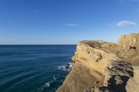 two people stand on the edge of a cliff next to the ocean in a bright blue sky