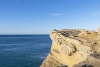 two people stand on the edge of a cliff next to the ocean in a bright blue sky