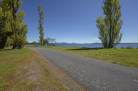 a dirt path beside a wide grassy field and a body of water with a line of trees to it