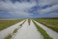 a long dirt road with some fencing around it in the middle of nowhere, near to a beach and grassy landscape