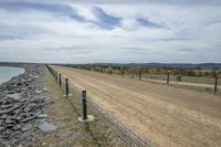 a dirt road and fence with trees in the background on a cloudy day near a body of water