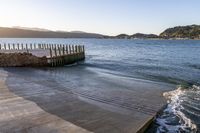 a wooden dock next to the ocean with waves hitting onto it at sunset, with hills in background