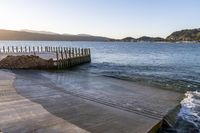 a wooden dock next to the ocean with waves hitting onto it at sunset, with hills in background