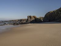 a sandy beach with rocks and clear skies above it by the water and rocks at the shore