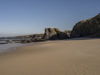 a sandy beach with rocks and clear skies above it by the water and rocks at the shore