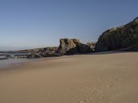 a sandy beach with rocks and clear skies above it by the water and rocks at the shore