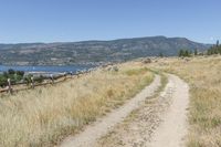 dirt path on the side of a grassy hill, with mountains in the background with lake behind and field with brown grass