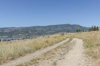 dirt path on the side of a grassy hill, with mountains in the background with lake behind and field with brown grass