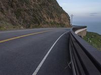 a curved road beside the ocean with rocky hills and a body of water in the background