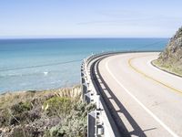 a highway near the coast with a road fence on each side and an ocean and hill in the background