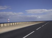 an empty road near a sandy shore in front of some water and sand with a few white clouds above