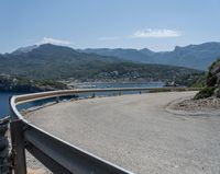 a curve on a highway near the ocean with mountains behind it and people walking by