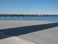 the shadow is cast on the asphalt near a large body of water and a bridge