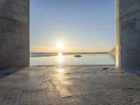 a sun shines behind two stone pillars overlooking a bay and a blue sky that has water