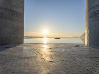 a sun shines behind two stone pillars overlooking a bay and a blue sky that has water