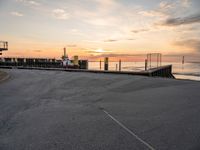 a man on a skateboard rides down the street by a pier at sunset in this photo