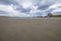 a picture of a beach with some very big rocks in the background, under a cloudy sky