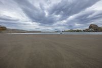 a picture of a beach with some very big rocks in the background, under a cloudy sky