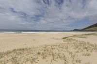 sandy beach with blue skies and waves crashing toward it by the ocean and hills in the distance