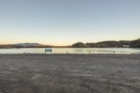 a bench sitting in an empty field near a body of water with mountains in the background