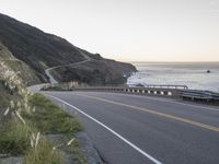 a curved highway next to an ocean at dusk with mountains in the background and the sun coming out over the mountains