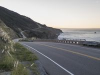 a curved highway next to an ocean at dusk with mountains in the background and the sun coming out over the mountains