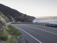 a curved highway next to an ocean at dusk with mountains in the background and the sun coming out over the mountains