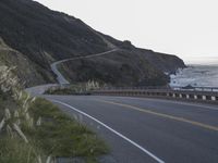 a curved highway next to an ocean at dusk with mountains in the background and the sun coming out over the mountains