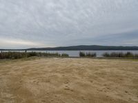 the sand area in front of a lake near a bench and bike rack with a few other picnic benches