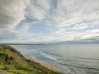 an empty bench sitting on a lush green hillside over looking the ocean and beach and hills