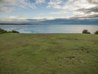 a large grassy field next to a body of water with clouds overhead the picture shows a man sitting at the edge of the grass hill overlooking an ocean