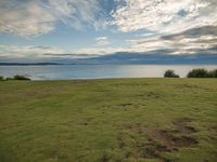 a large grassy field next to a body of water with clouds overhead the picture shows a man sitting at the edge of the grass hill overlooking an ocean