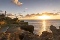 rocks and a lighthouse are at the ocean shore and sunset light shines through the clouds