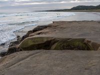 there are large rocks sitting on the edge of the water and a surfer is in the distance