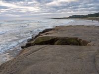 there are large rocks sitting on the edge of the water and a surfer is in the distance