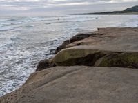 there are large rocks sitting on the edge of the water and a surfer is in the distance