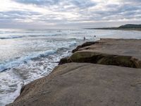 there are large rocks sitting on the edge of the water and a surfer is in the distance