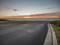 an empty road on a coast near the ocean in front of a sunset sky above the water