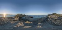 the sun rises over rocks at the beach with the ocean behind them as a fisheye lens