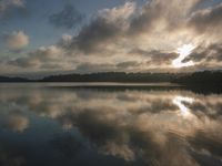 a large body of water with trees in the background and clouds above it and sun reflecting on the water