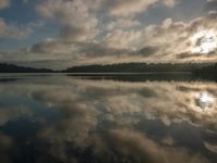 a large body of water with trees in the background and clouds above it and sun reflecting on the water