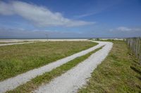 a dirt road that leads towards the water on the beach and onto a fence and grass field