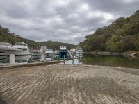 several small boats docked on water next to a pier or dock under cloudy skies and a tree covered hill