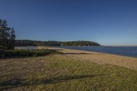 a grassy field next to the water with a small beach in the distance with no people at the end